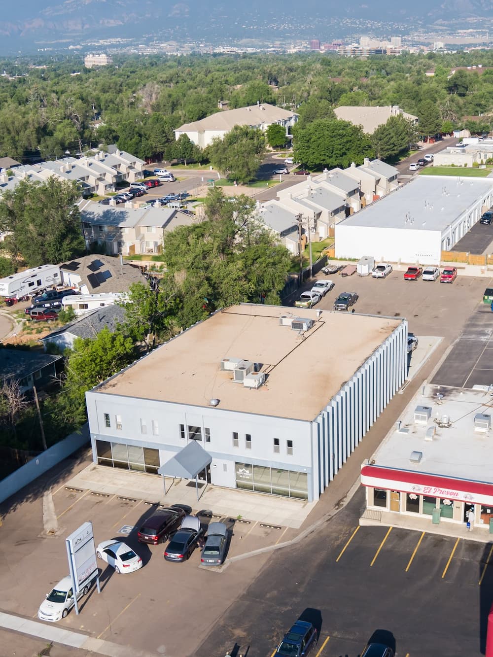 Aerial view of a two-story light blue commercial building with a flat roof, surrounded by a parking lot and neighboring residential and commercial buildings.