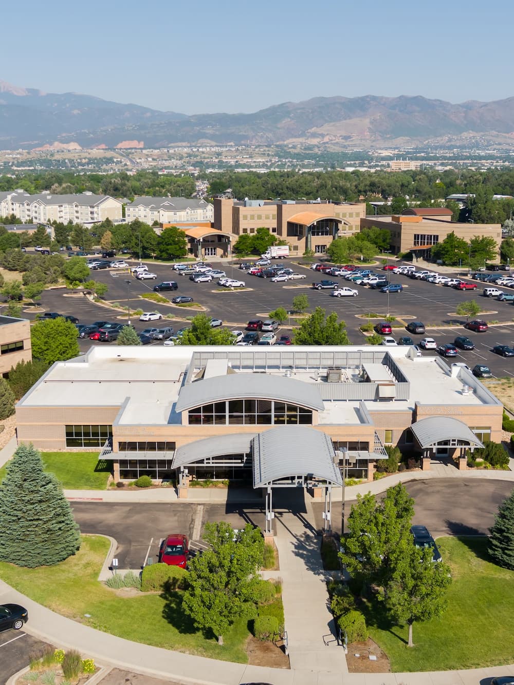 Aerial view of a complex of tan-colored office buildings with large parking lots, surrounded by trees with a mountain range in the distance.