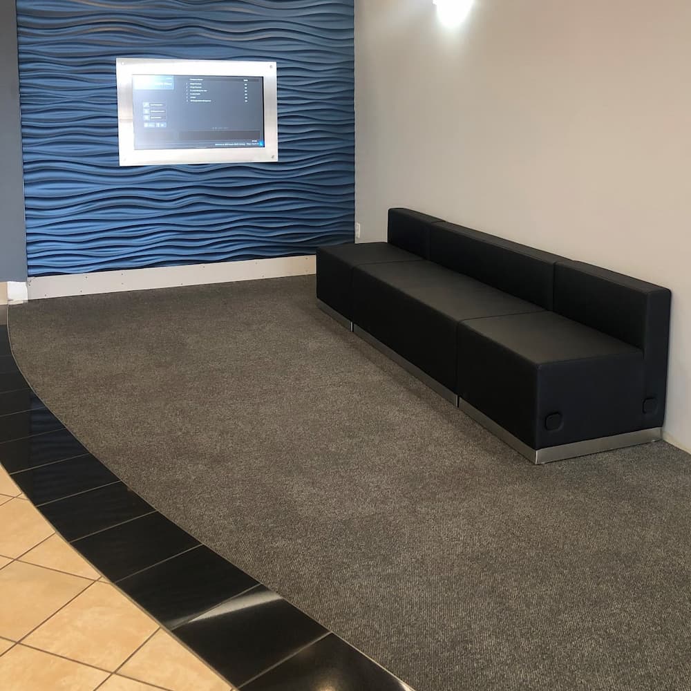 A black modular waiting area bench sits on gray carpet in front of a blue, wave-textured wall featuring a wall-mounted digital display screen.