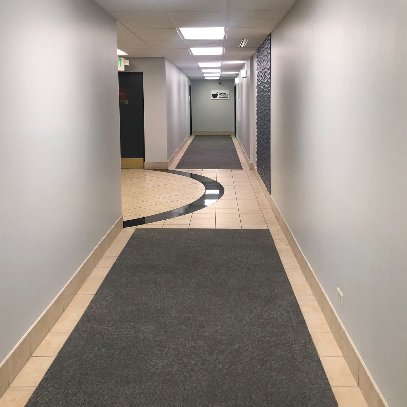 An empty office building hallway with grey carpet runners, tiled floors, and a sign for a pharmacy.