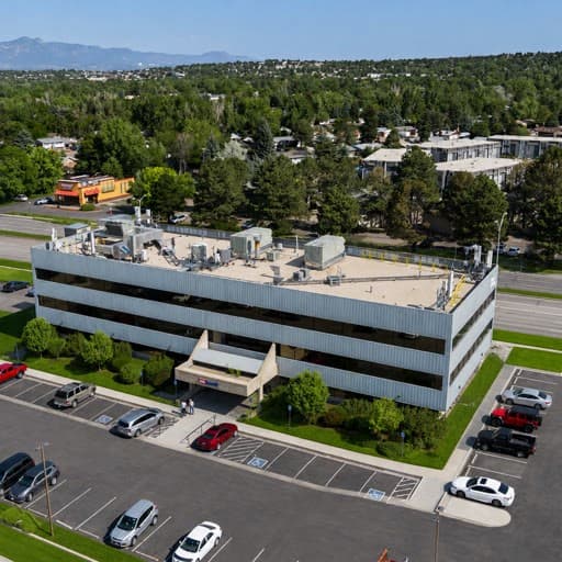 An aerial view of a multi-story office building with a flat roof featuring several HVAC units, surrounded by a parking lot with various cars.