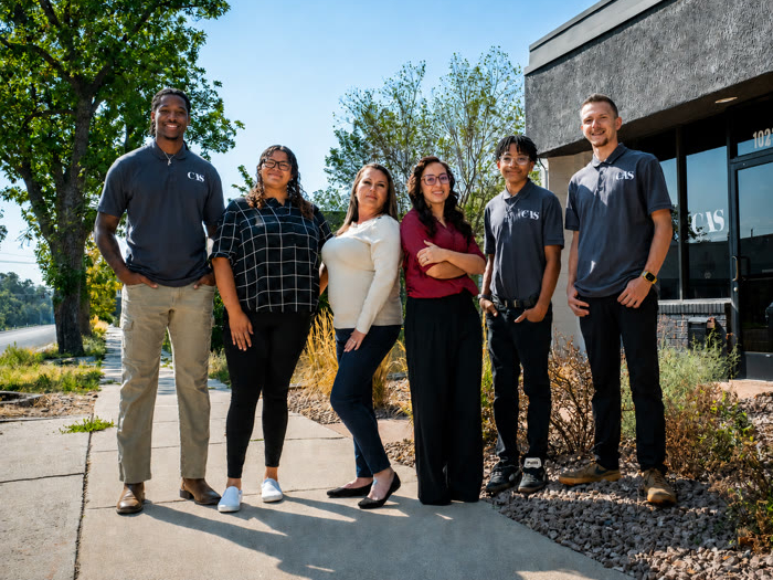 Five employees in casual attire, some wearing shirts with the CAS logo, stand in a line on a sidewalk outside an office building.