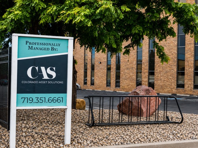 A sign for Colorado Asset Solutions with the phone number 719-351-6661, standing in front of a brick building with a bike rack and large rock.
