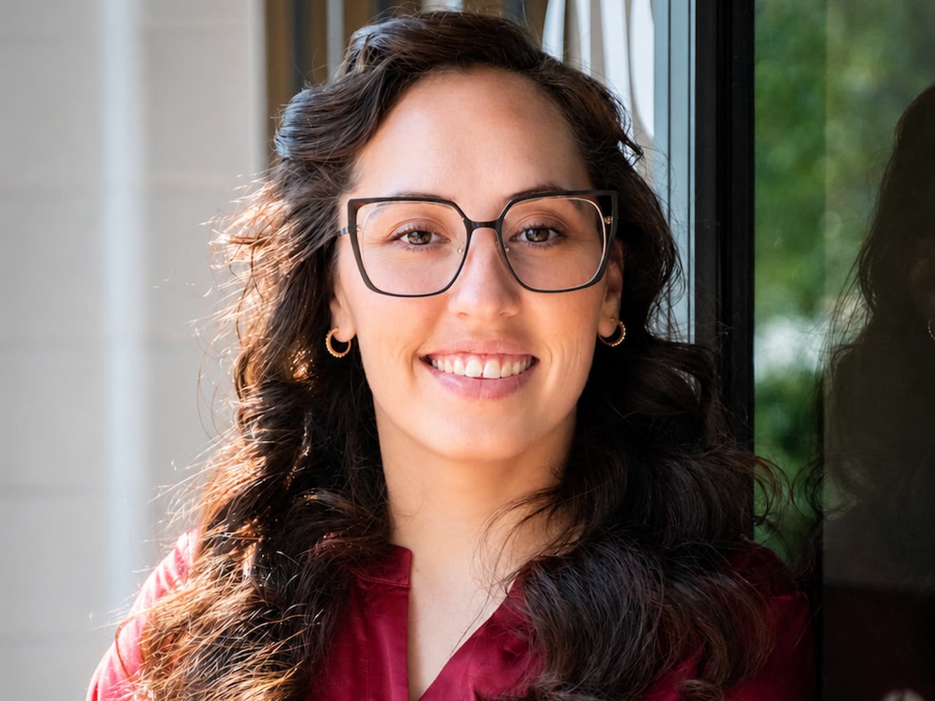 A smiling woman with dark wavy hair wearing black-rimmed glasses and small gold hoop earrings.