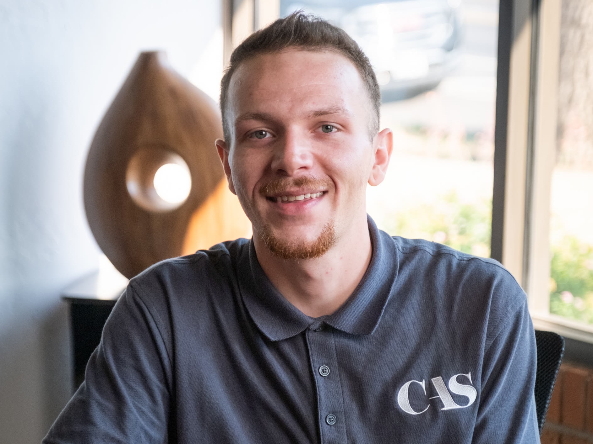 A smiling man with a short beard wearing a grey polo shirt with “CAS” embroidered on the chest.