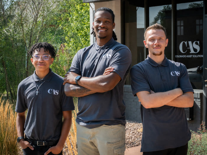 Three employees wearing grey Colorado Asset Solution polo shirts standing outside the company office.