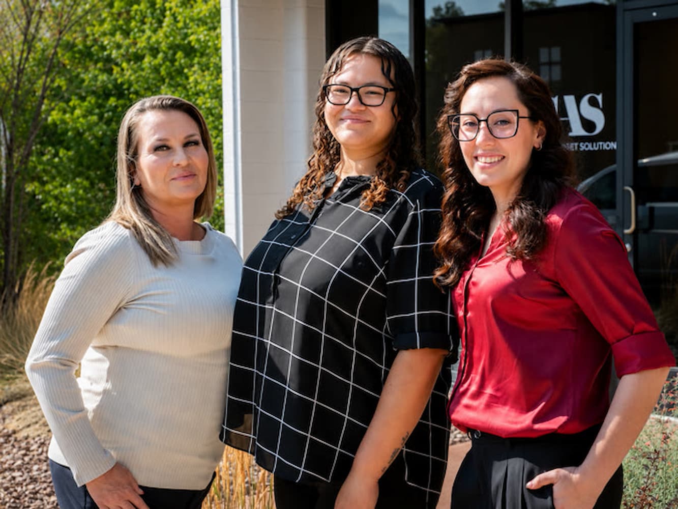 Three women standing together outdoors in front of a building entrance.