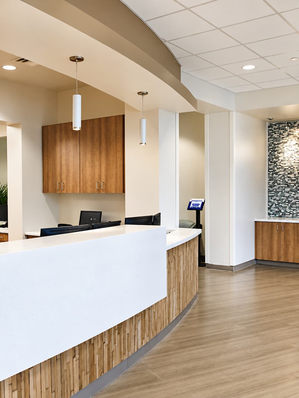 A curved reception desk in a modern office with light wood paneling, white counters, and a mosaic tile wall in the background.